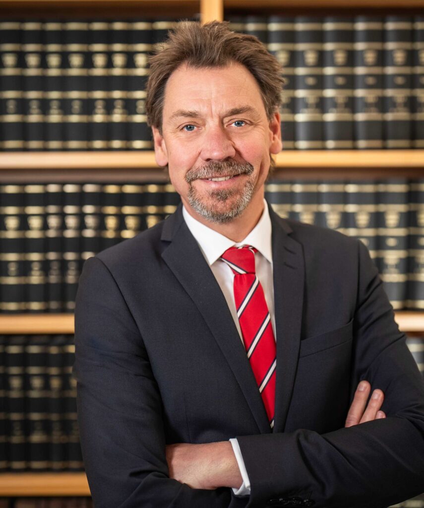 Porträt eines erfahrenen Rechtsanwalts und Wirtschaftsprüfers im dunklen Anzug mit rot-gestreifter Krawatte vor einem Bücherregal – Experte für Rechtsberatung und Wirtschaftsprüfung im Norden