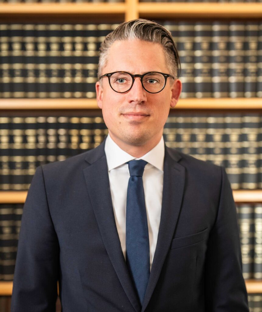 Porträt eines Rechtsanwalts und Wirtschaftsprüfers im dunklen Anzug mit blauer Krawatte und Brille vor einem Bücherregal – Fachmann für Rechtsberatung und Wirtschaftsprüfung im Norden