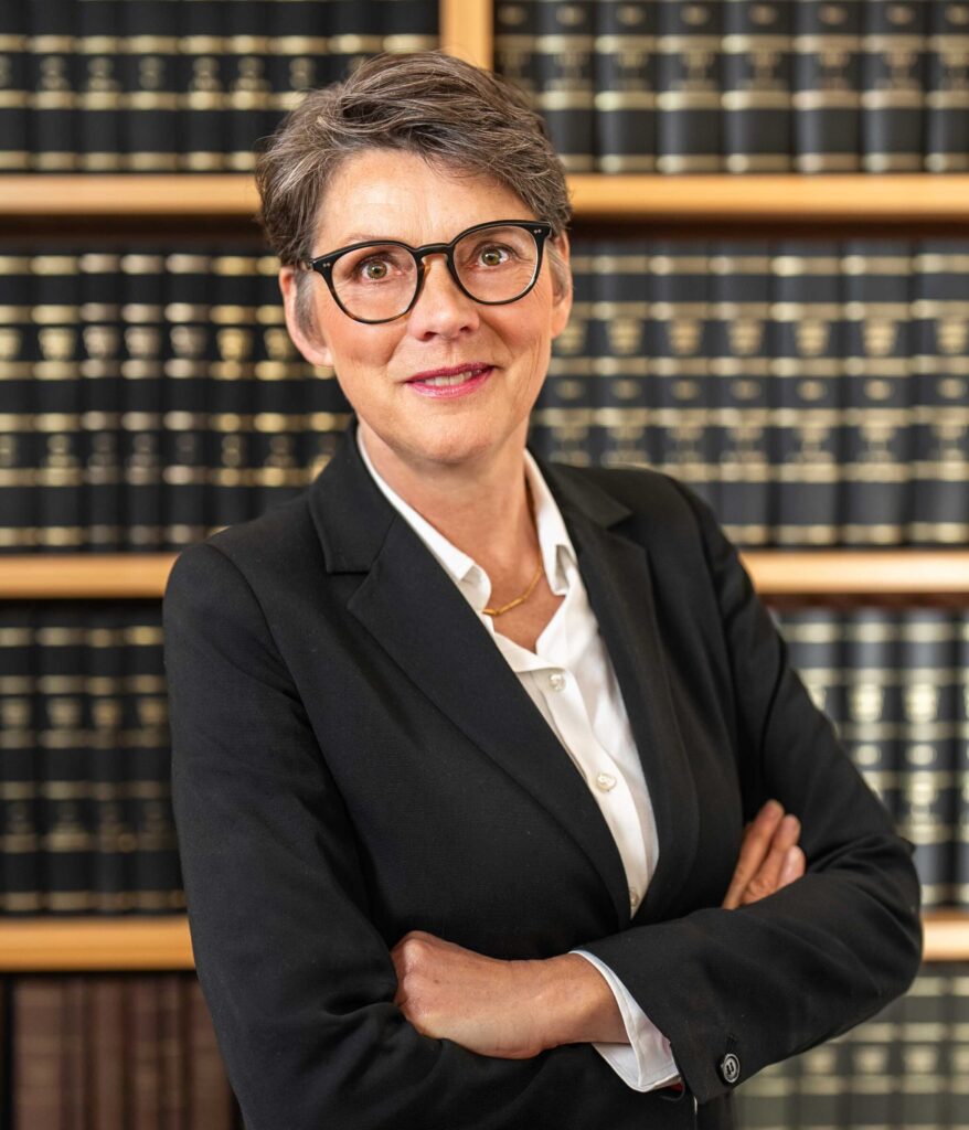 Porträt einer erfahrenen Rechtsanwältin und Wirtschaftsprüferin im schwarzen Blazer mit Brille und verschränkten Armen vor einem Bücherregal – Spezialistin für Rechtsberatung und Wirtschaftsprüfung im Norden